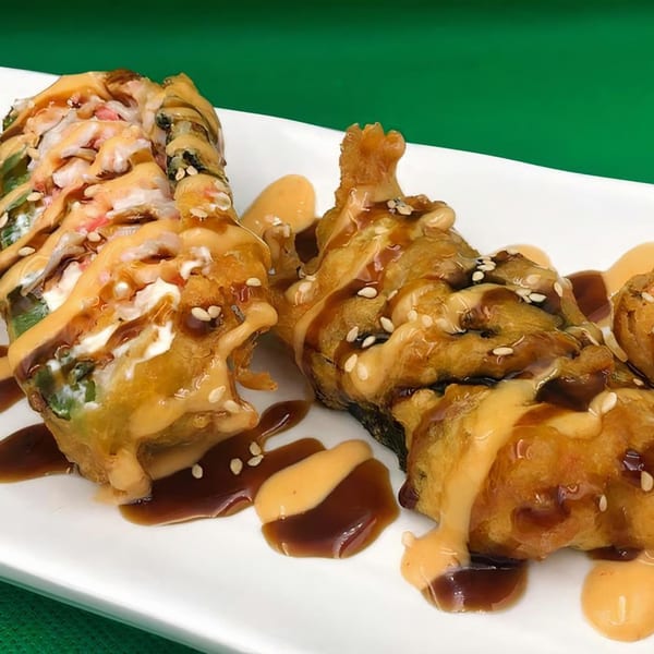 Two deep-fried sushi rolls drizzled with creamy sauce, dark sauce, and sesame seeds on a white plate.