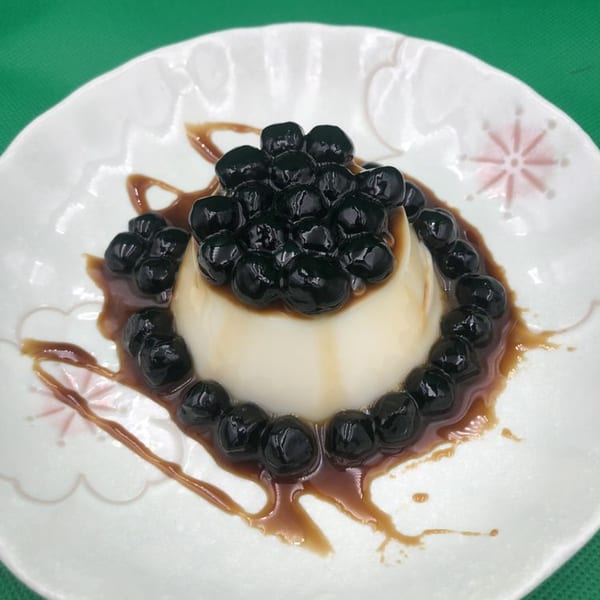 Panna cotta dessert topped with dark boba pearls and caramel drizzle on a white plate.
