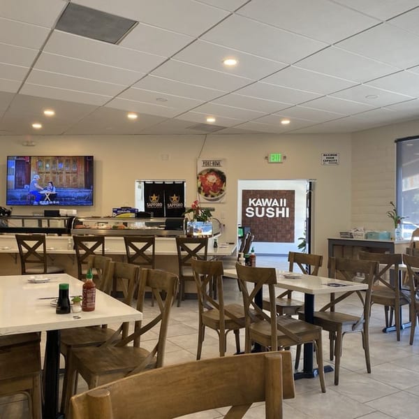 Interior of Kawaii Sushi restaurant with empty tables, chairs, a sushi bar, and a TV playing.
